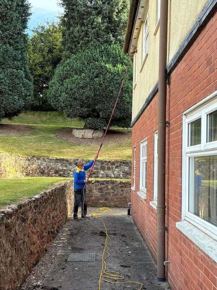 Window cleaner using a long pole on upper windows beside a garden